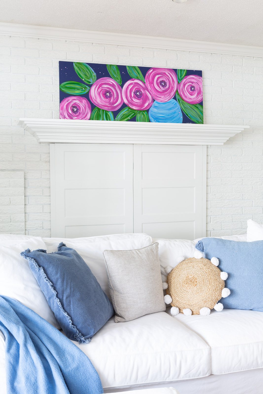 white living room with a DIY art canvas of large pink flowers on a blue background leaning on fireplace mantel.