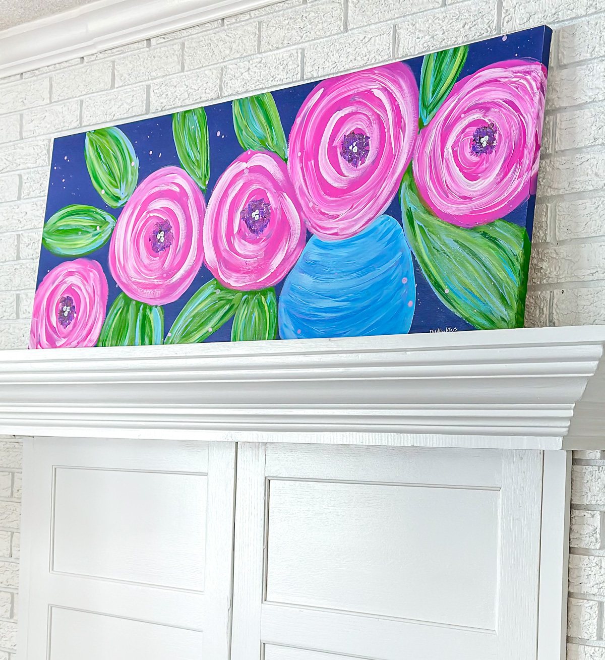 DIY wall art painting of big pink flowers on a dark blue background placed on a living room mantel.