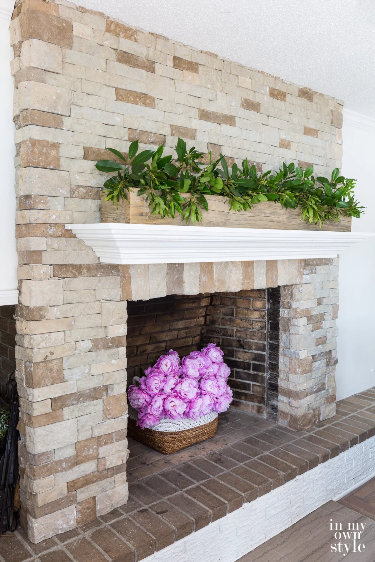Stone fireplace with white mantel. Pink flowers in fireplace. Greenery clippings in wood planter on top of mantel.