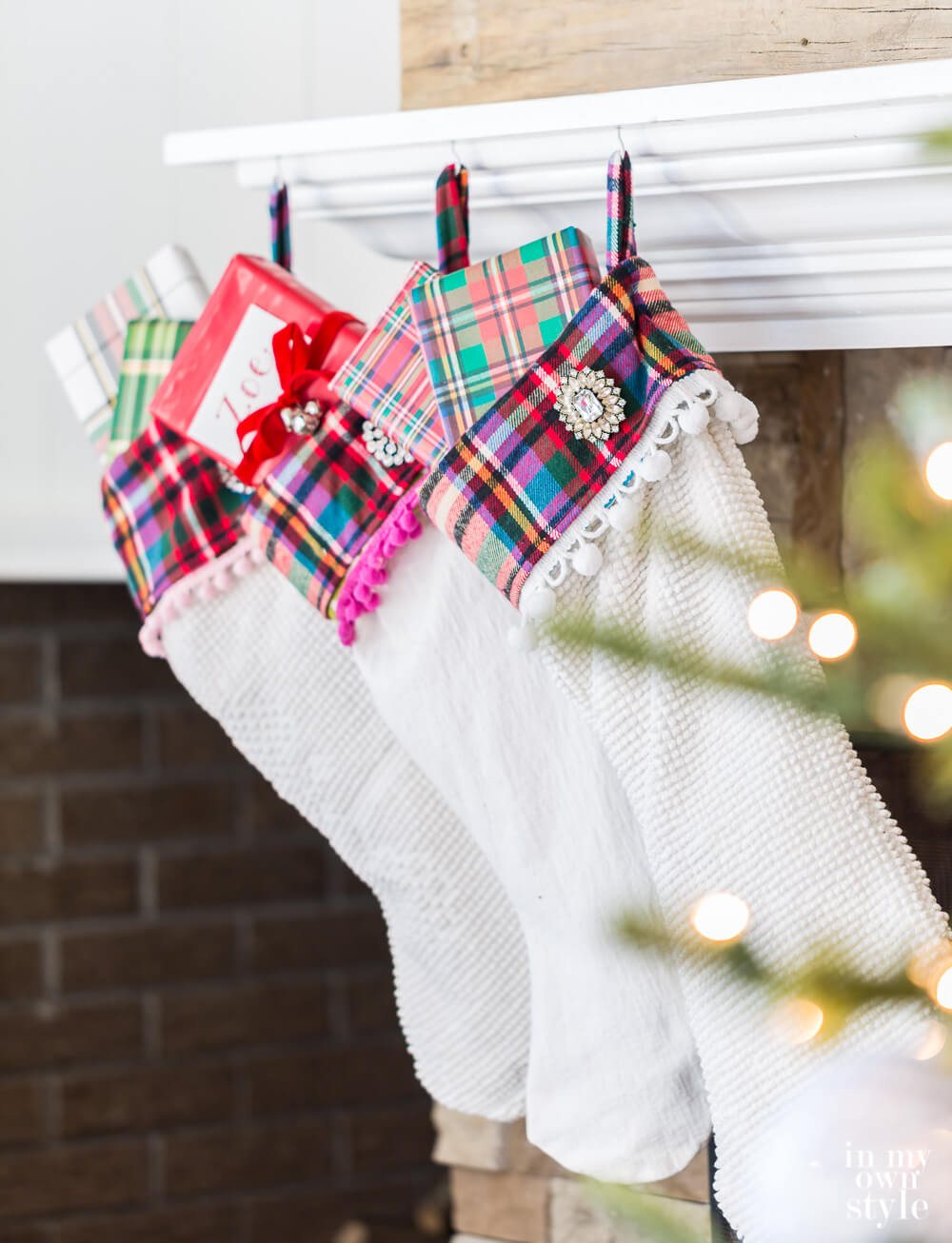 3 white Christmas stockings with plaid cuffs and pom-poms hanging from a fireplace mantel.