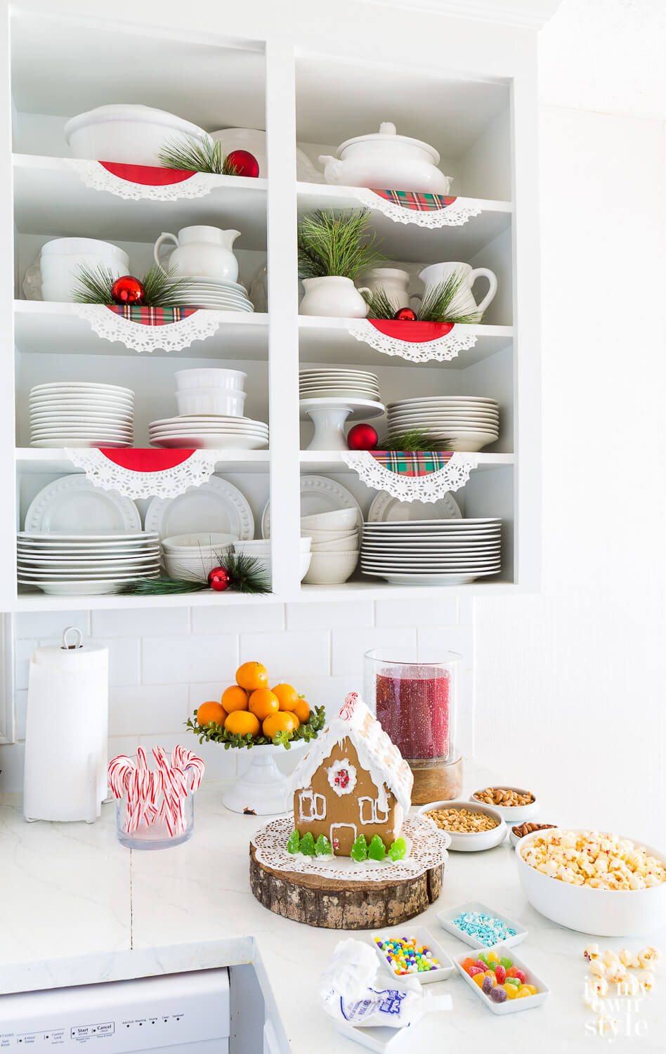 Open white shelves in kitchen. Red and plaid paper doilies line each shelf. Red ornaments and pine sprigs tucked in randomly.