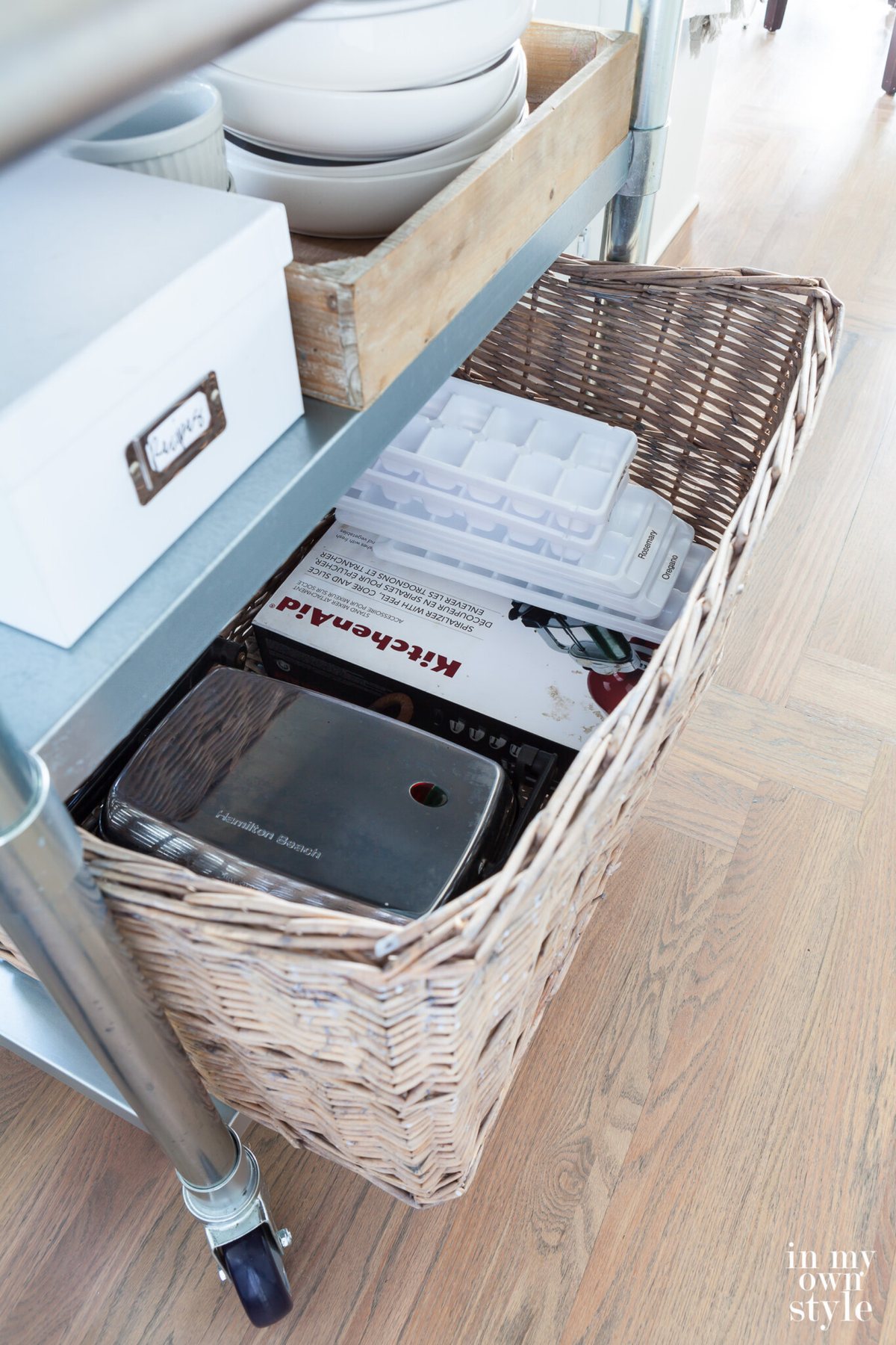 A large wicker basket on a metal rolling kitchen island filled with least used kitchen tools.