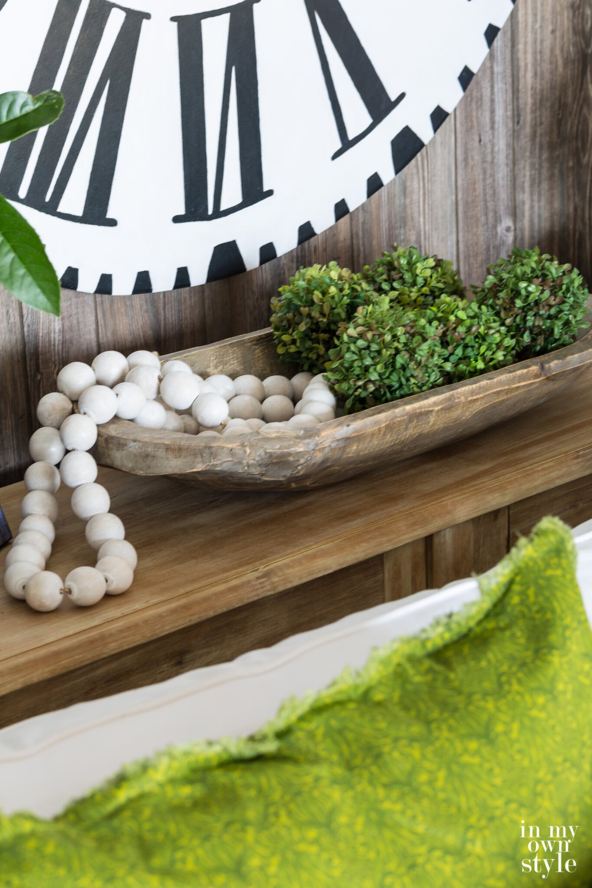 Dough bowl on sofa table filled with moss globes and wood beads. white clock with black Roman numerals on the wall.