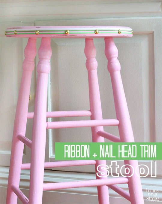 Pink painted stool with ribbon and nail head trim around the edge of the seat.