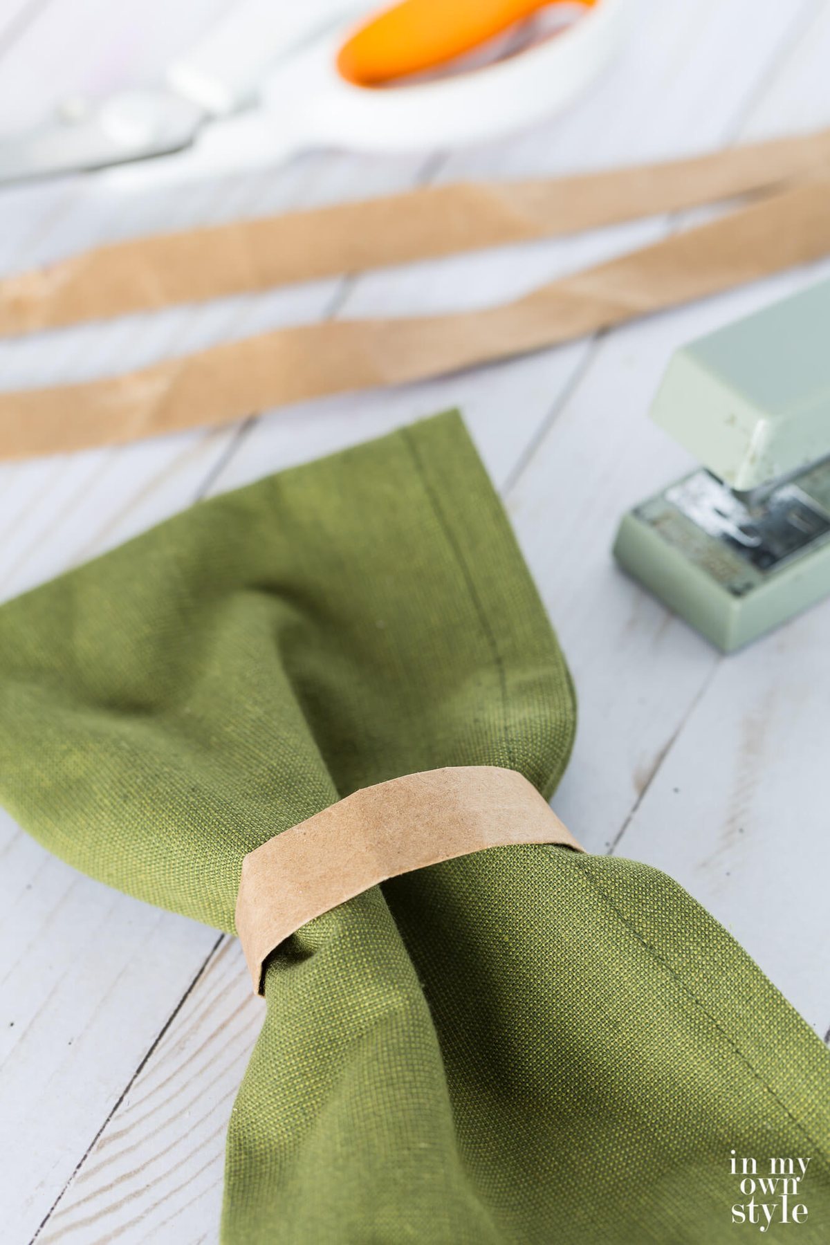 Brown paper bag handle napkin ring on dark sage green napkin. Stapler and scissors in the background.