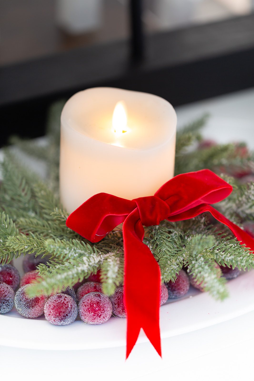 close up of a white candle on a platter with a Christmas wreath around it and sugared cranberries.
