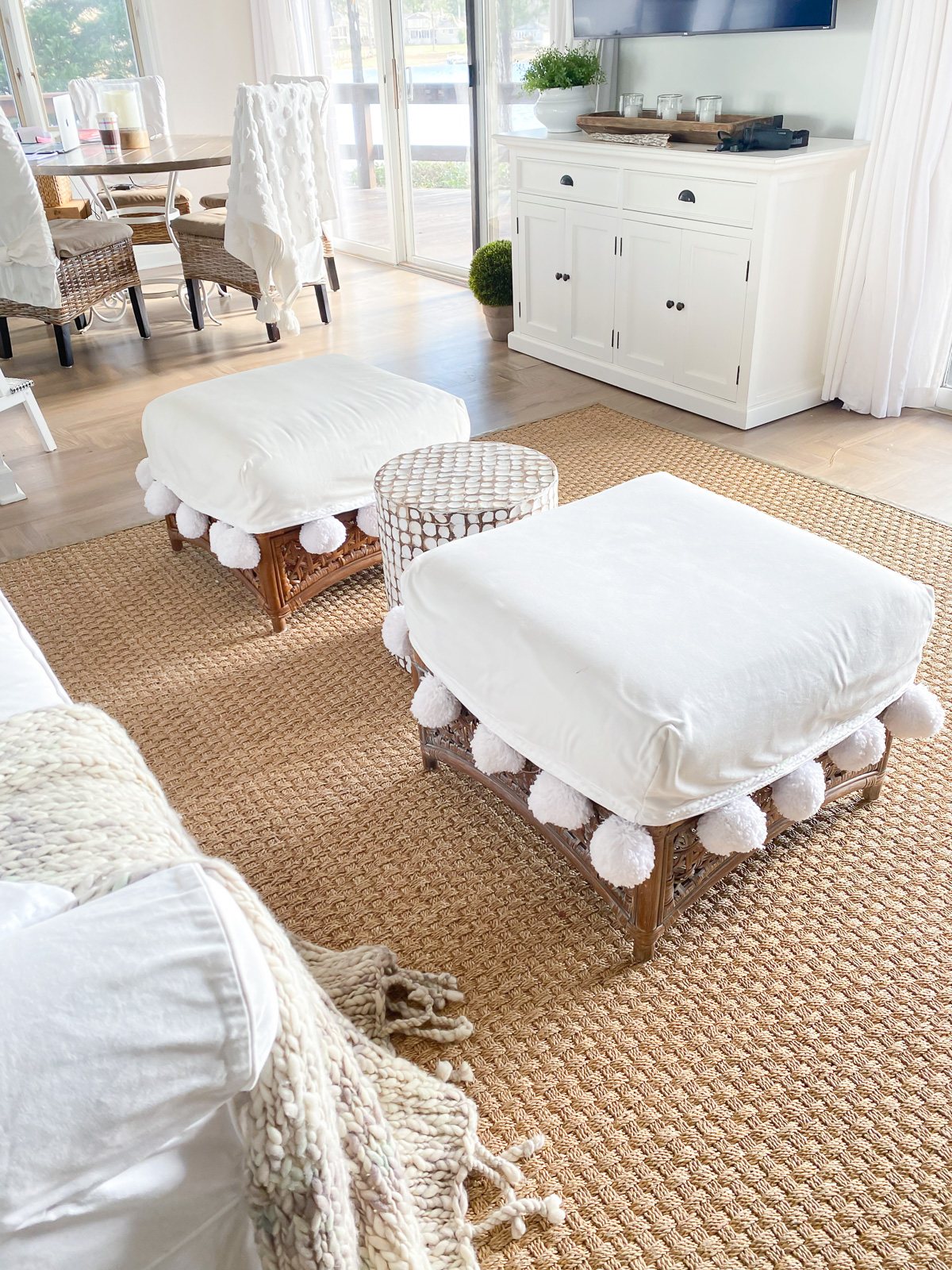 two square rattan ottomans with large pom-pom trimmed cushions in a living room.