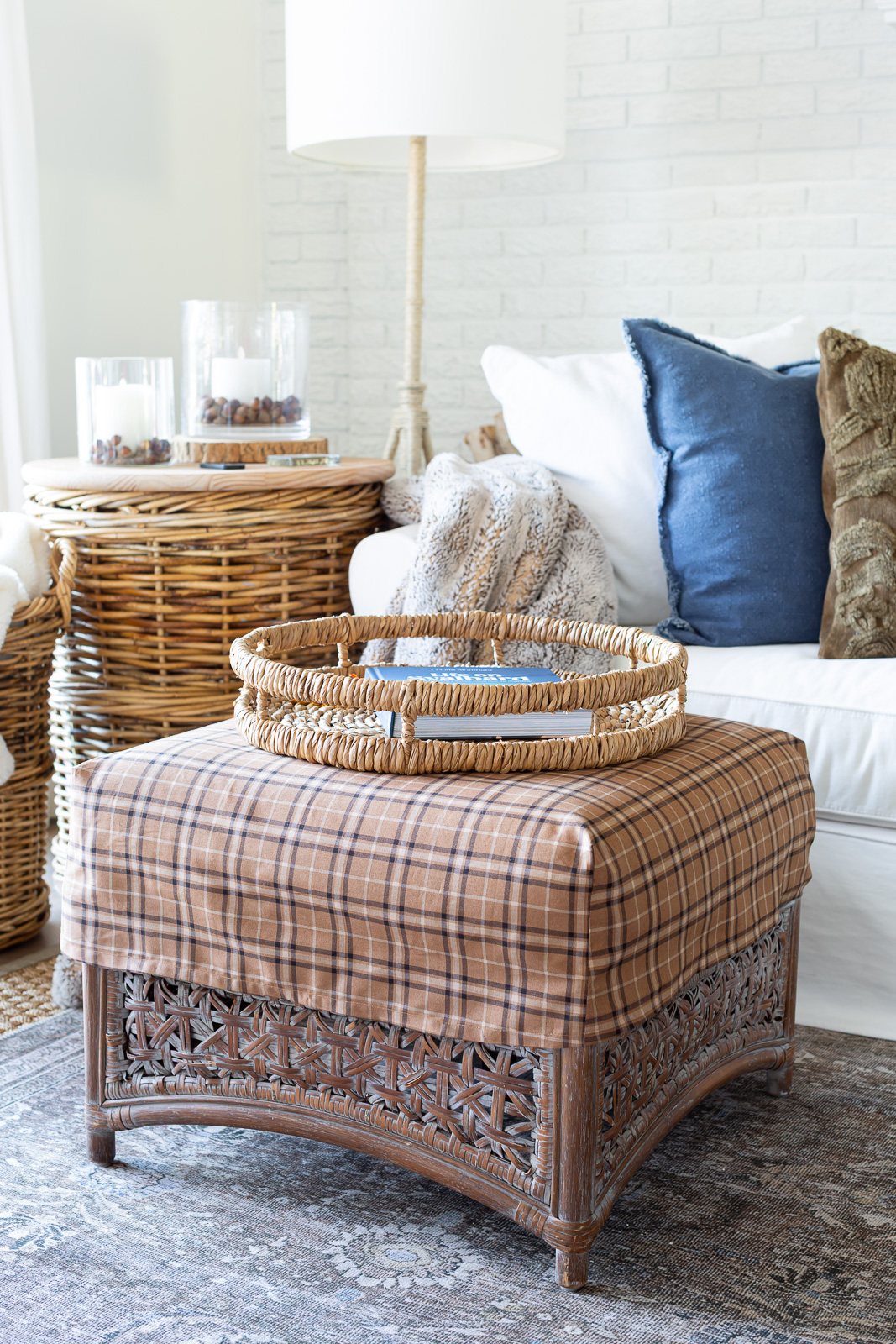 a square rattan ottoman with a brown plaid slipcover on it in from of a white sofa with layers of fall decor on it.