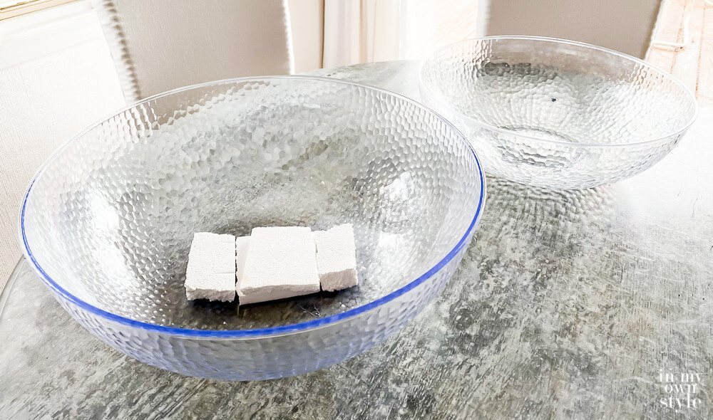 Two large clear plastic bowls on table. One bowl has 3 small pieces of white Styrofoam inside.