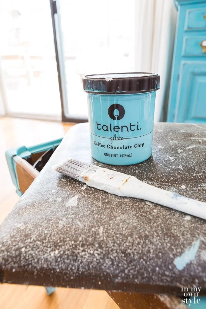 An plastic gelato container being used to hold turquoise chalk paint. It is placed on the unfinished stool with a paint brush.