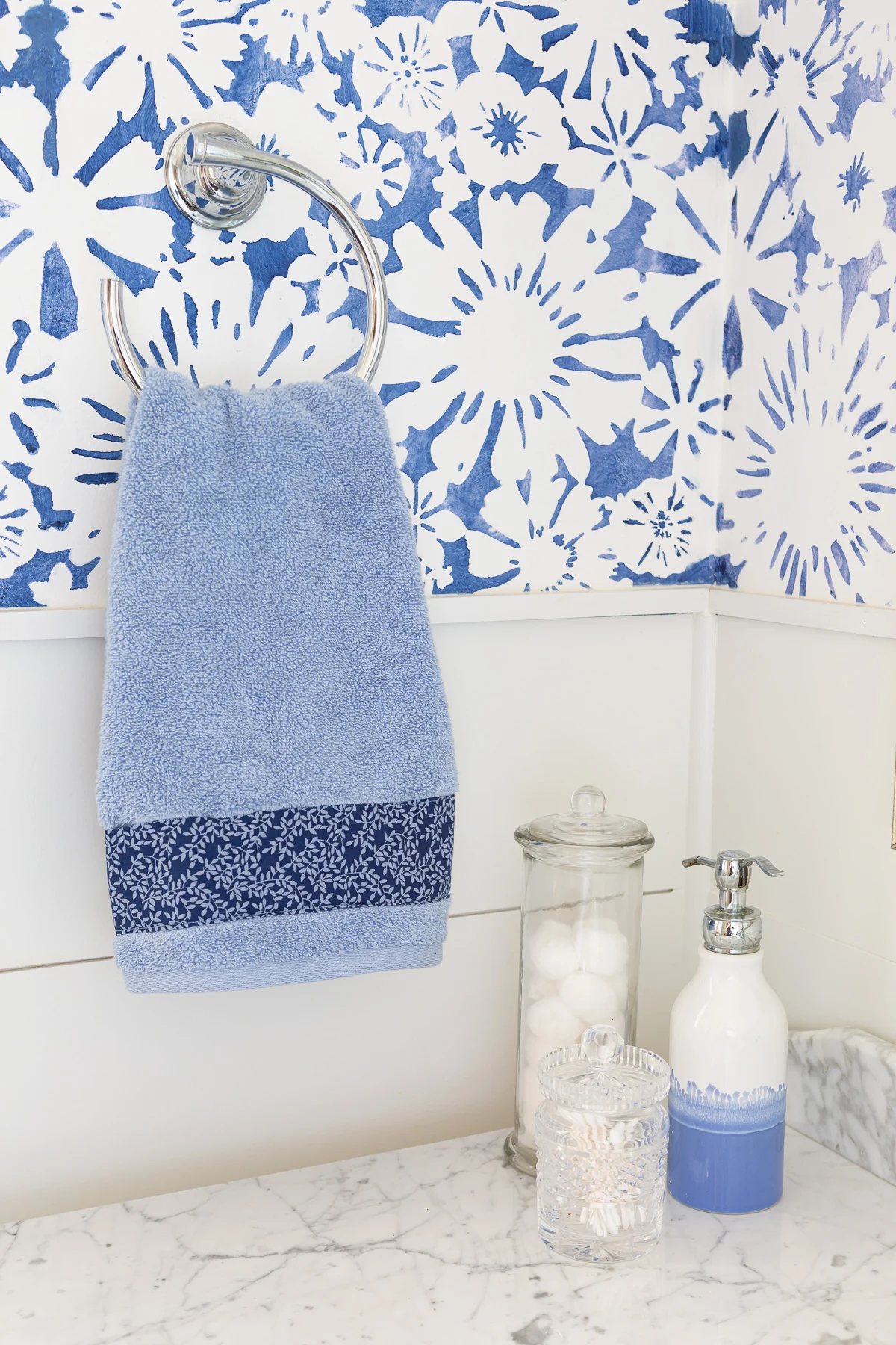 sky blue hand towel hanging from a towel ring. Decorative fabric was added to the bottom edge of the towel to embellish it.