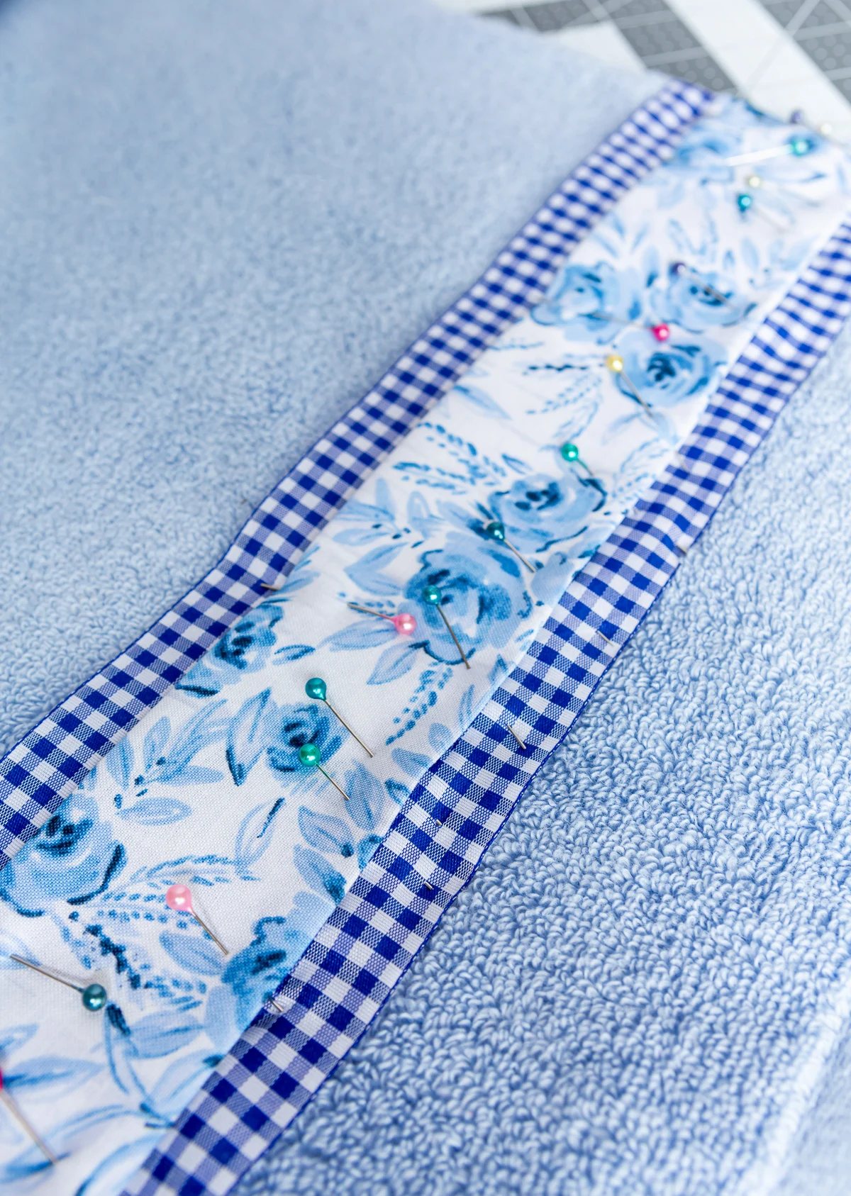 Blue flower fabric and gingham ribbon being pinned onto a bath towel before sewing.