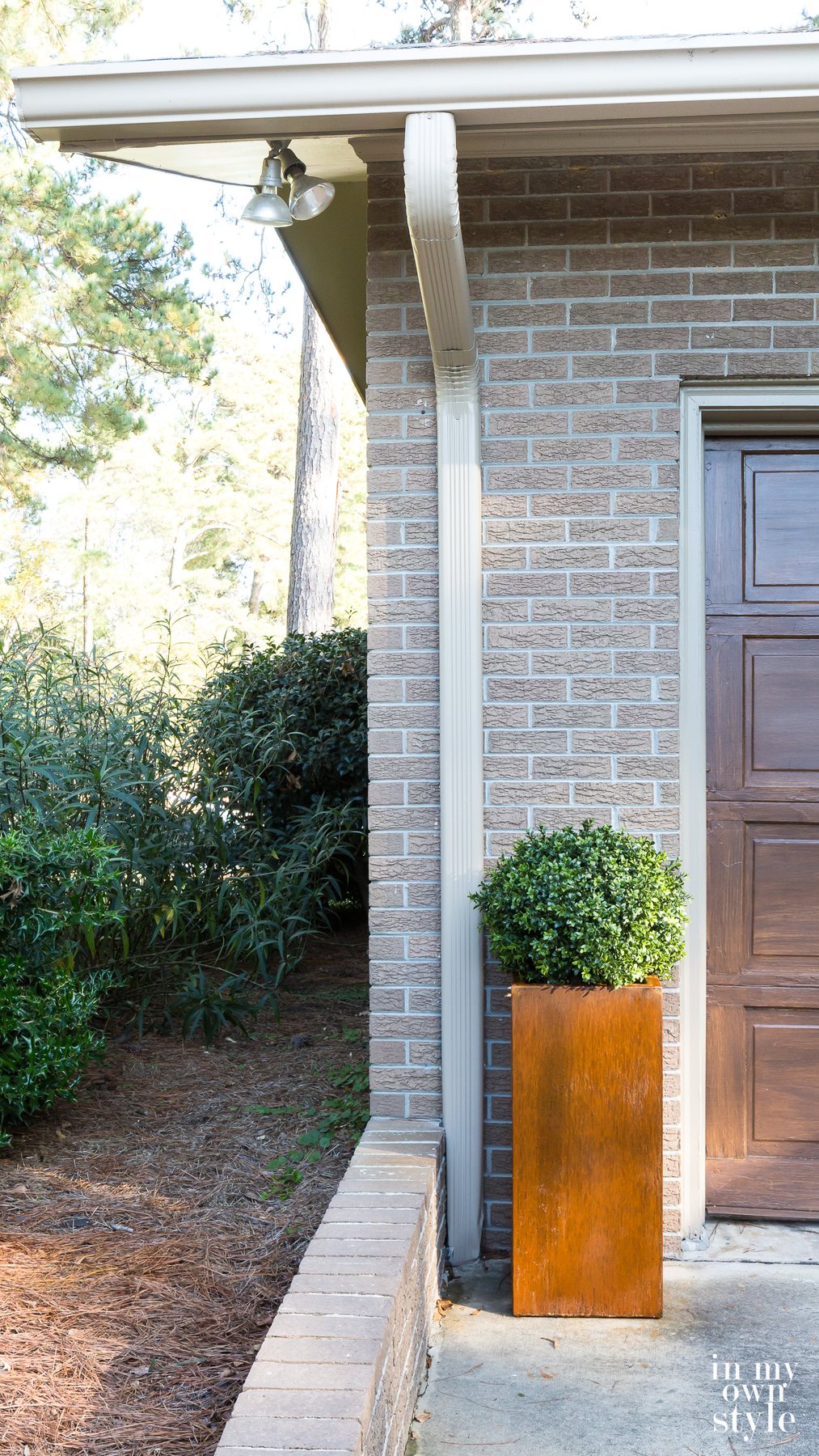 Rusted Corten Steel tall planter with boxwood placed on a driveway next to a garage door.
