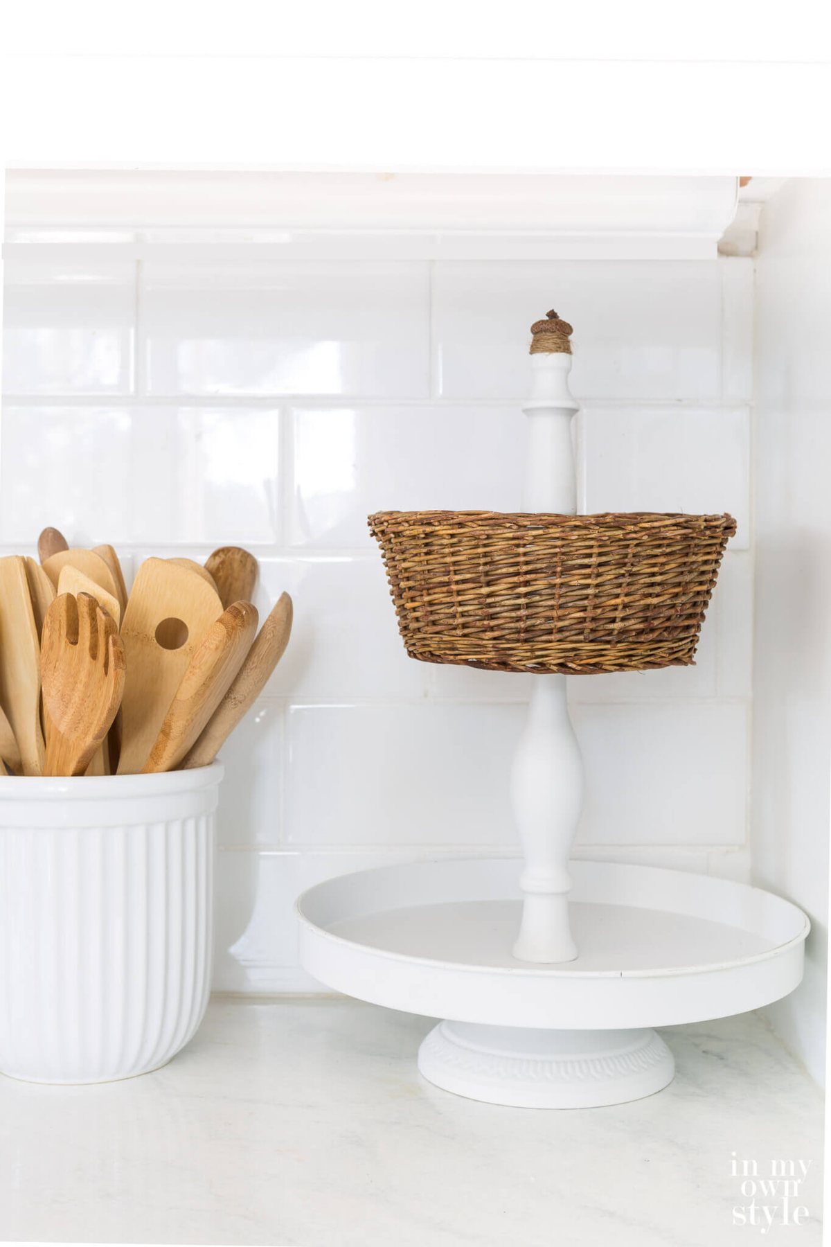 White metal 2 tiered stand with wicker basket on kitchen counter.