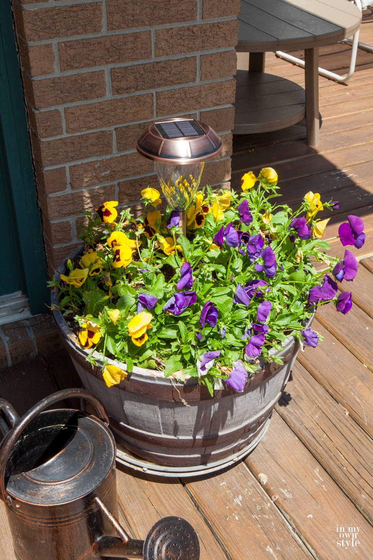 Copper solar light in pansy filled planter. Watering can next to it. Another way to add outdoor deck lighting in an easy way.