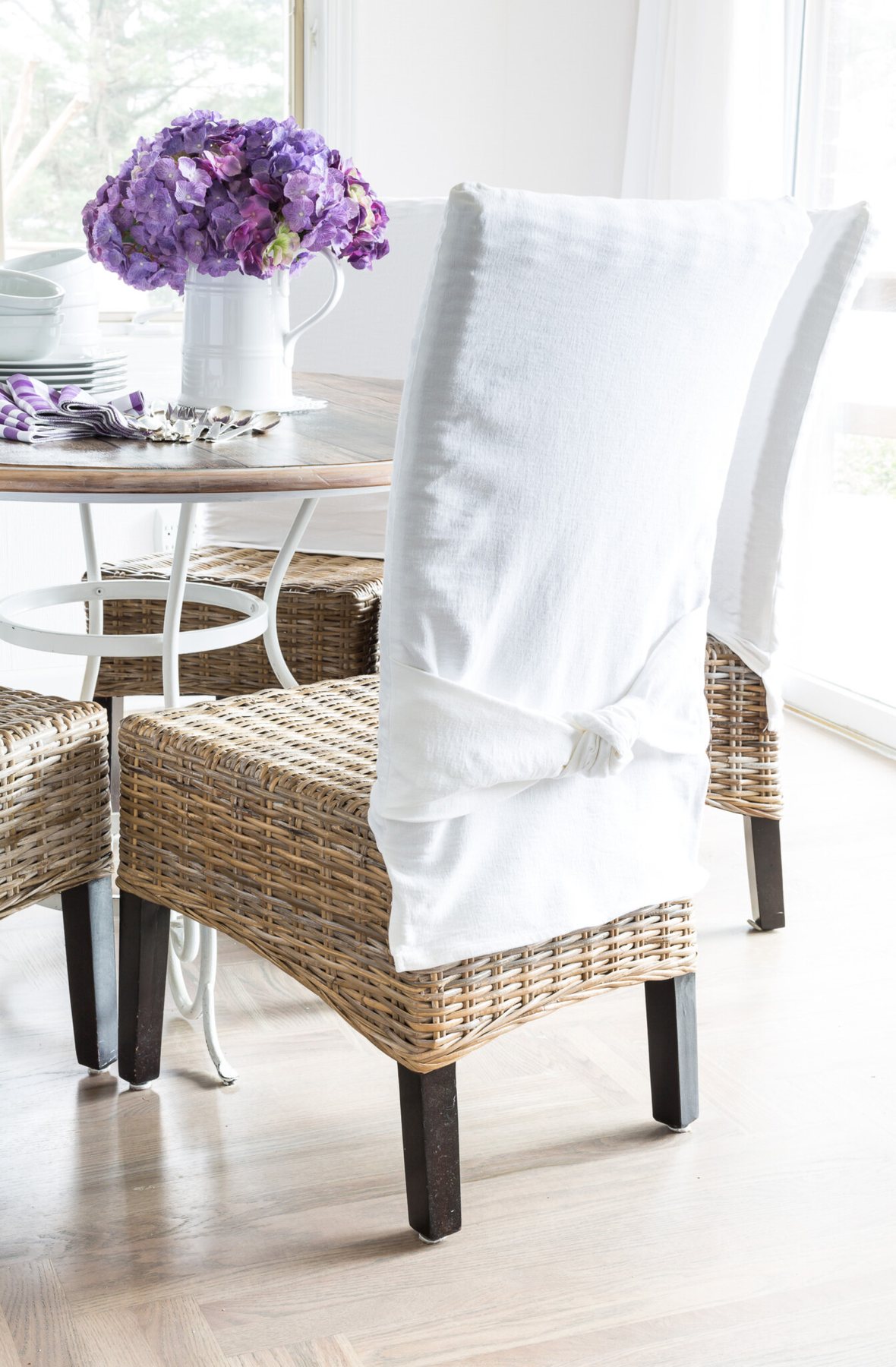 Wicker chairs around a round dining room table. Chairs have white slipcovers with a tied knot detail in the chair backs.