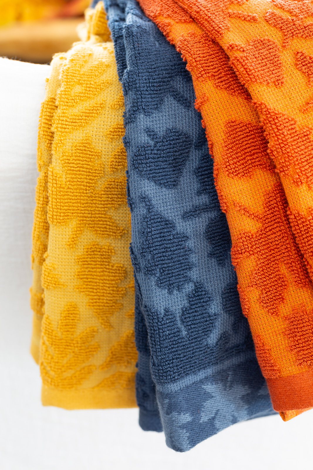 3 fall hued dishtowels with a textured leaves draped over a chair.