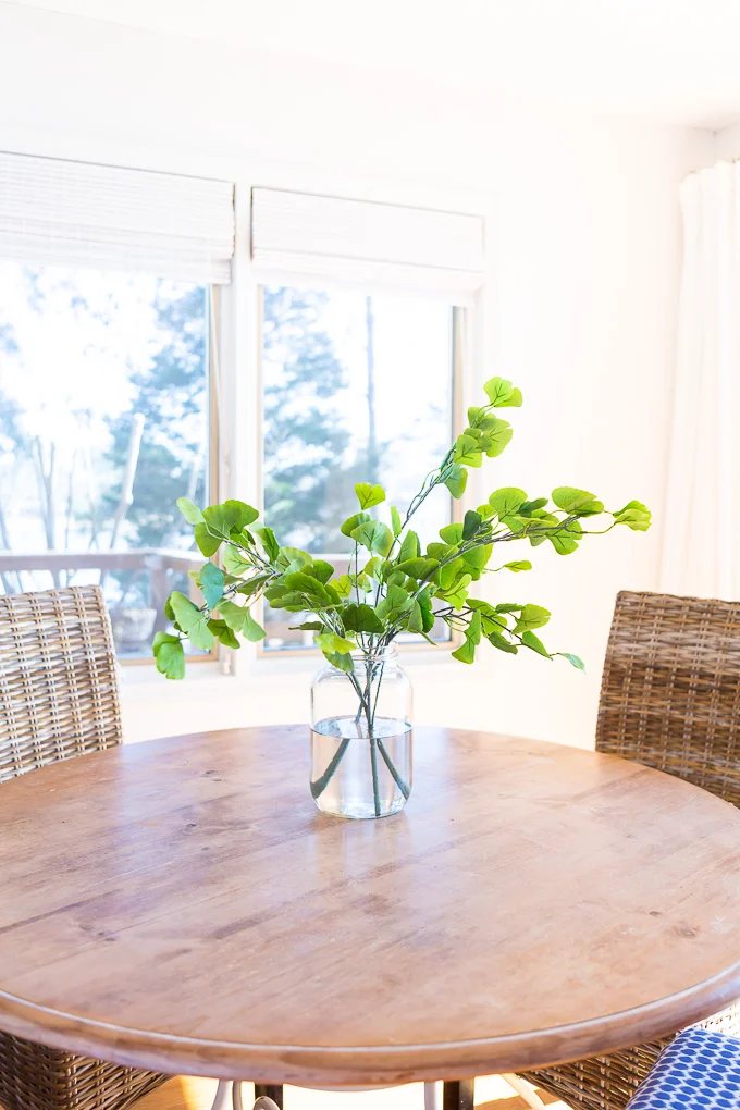 Large glass vase filled with water and fake house plant that is sitting on a round wood table.