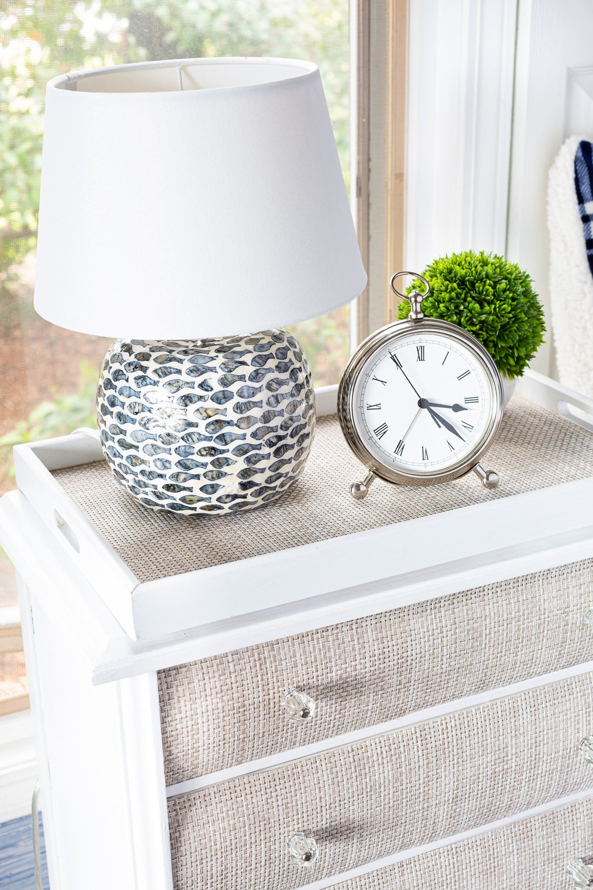 A night table with a table lamp on a round wood riser, silver alarm clock and a small plant.