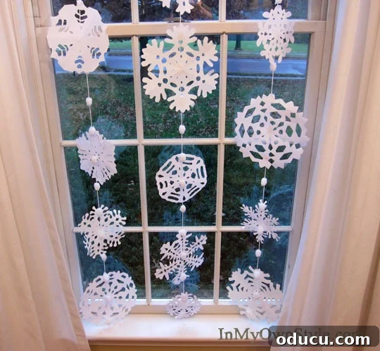 DIY Paper Snowflake Window Displays 16 A beautiful arrangement of paper snowflakes hanging in a window, adorned with multiple pom-poms.