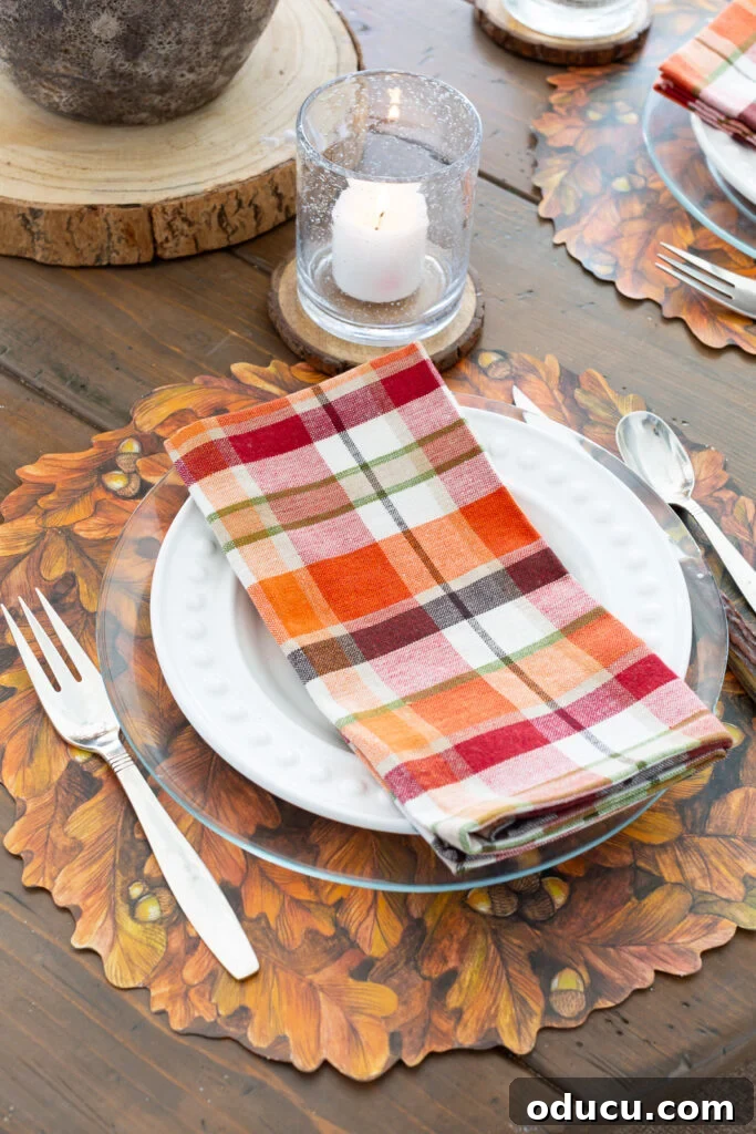 Thanksgiving's Functional and Festive Focal Point 7 Thanksgiving table setting featuring a Hester & Cook fall leaves paper placemat, a plaid napkin on a clear glass plate, and elegant flatware.