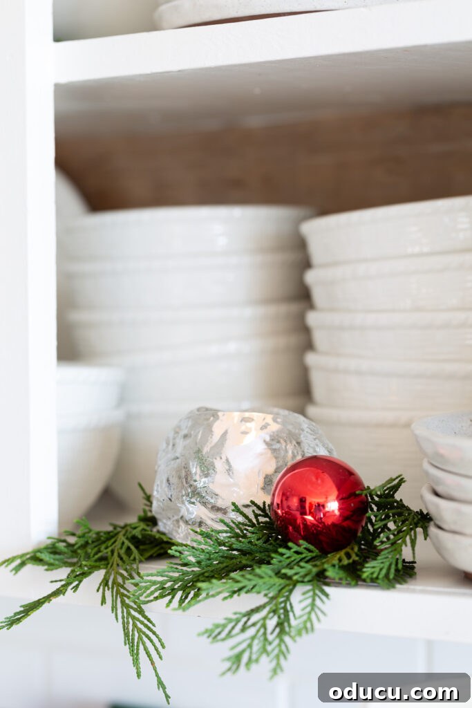 My Christmas Home: Timeless Memories, Not Trends 11 Kosta Boda snowball votive holder on an open kitchen shelf with greenery around it and a red ball ornament.