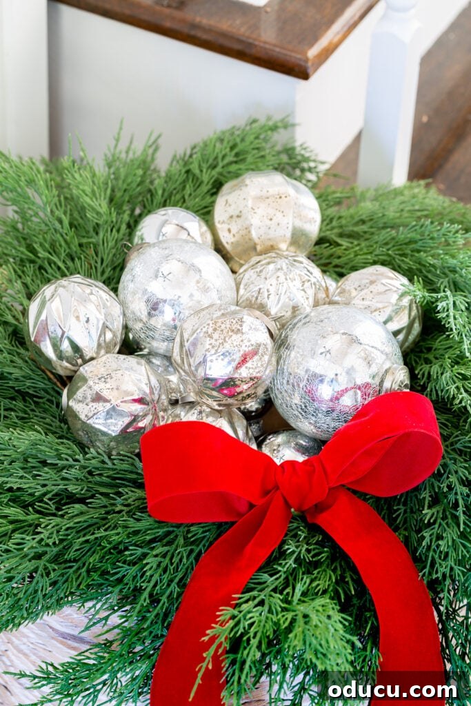 The Wreath Revelation You Need This Christmas 7 A collection of gleaming silver Christmas ornaments is nestled artfully within a vibrant greenery wreath, perched on a large woven floor basket, accented with a rich red velvet bow.
