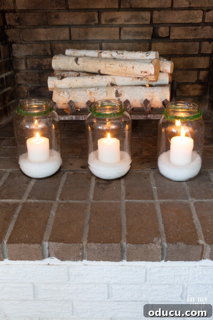 3 large glass jars filled with fake snow and white candles in a fireplace.
