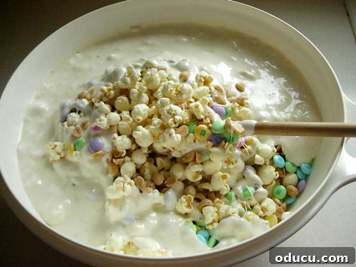 Popcorn-Cake-recipe getting mixed Hands mixing the warm marshmallow mixture into the popcorn, peanut, and M&M's in a large bowl, ensuring everything is thoroughly coated.