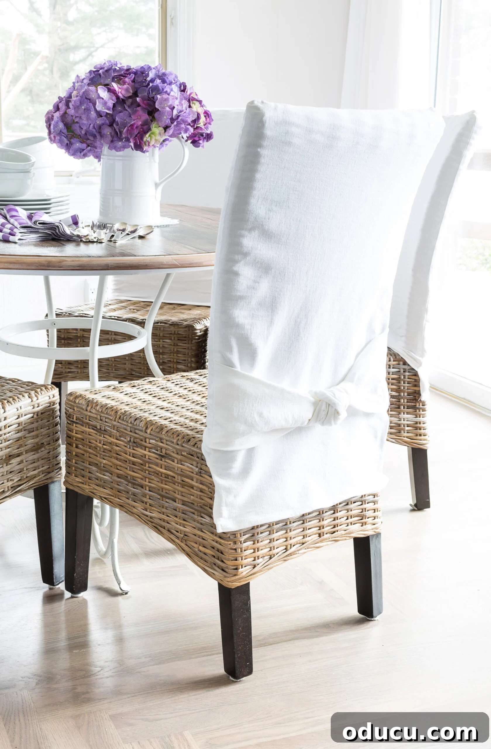 Transform Your Dining Chairs with Easy DIY Slipcovers 12 Wicker chairs around a round dining room table. Chairs have white slipcovers with a tied knot detail in the chair backs. A charming dining area featuring custom slipcovers, exemplifying style and comfort.