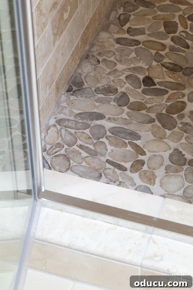 Luxury Master Shower Transformation 13 Close-up view of the new stone tile shower floor, featuring grey pebble mosaics, with the clear glass door swung open, revealing the spacious interior.