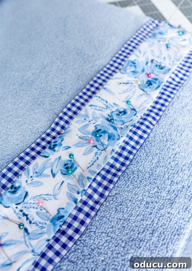 Elevate Your Bathroom With Towel Decor 12 A charming blue flower fabric and coordinating gingham ribbon being meticulously pinned onto a plain bath towel, preparing it for the sewing process.