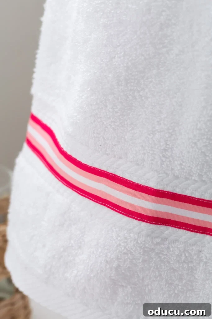 Elevate Your Bathroom With Towel Decor 6 A detailed view of a plain white bath towel, transformed with a neat stripe of pink and white striped grosgrain ribbon meticulously sewn onto its surface.