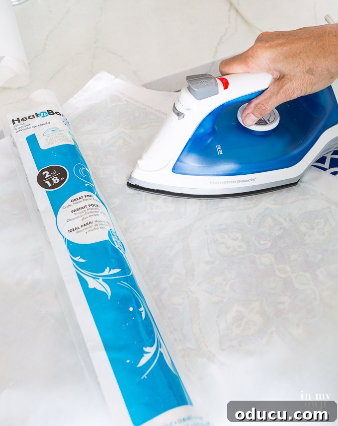 From Neglected Stool to Elegant Ottoman 11 Using a dry iron to press Heat n Bond vinyl onto fabric, demonstrating proper application technique.