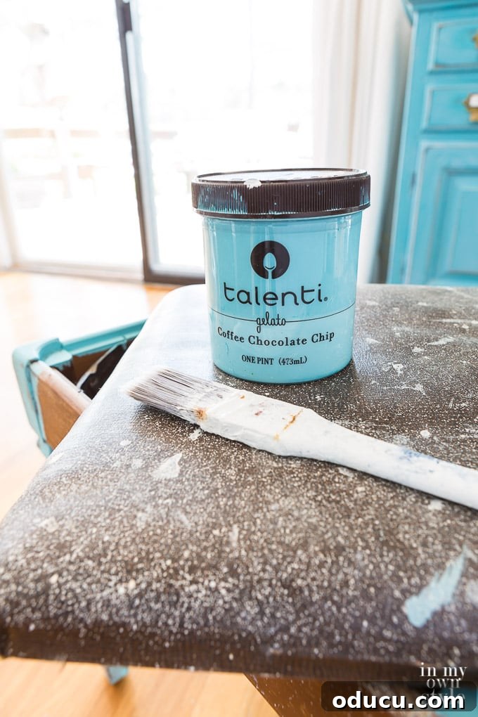 From Neglected Stool to Elegant Ottoman 4 A plastic gelato container holding turquoise chalk paint, a paintbrush, and the unfinished stool ready for painting.