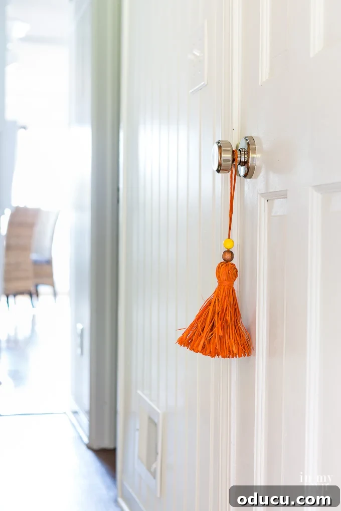 Handmade Raffia Tassel: A Perfect Fall Home Decor Accent A vibrant orange raffia tassel with a yellow bead, gracefully hanging from a doorknob, showcasing a handmade fall decor accent.