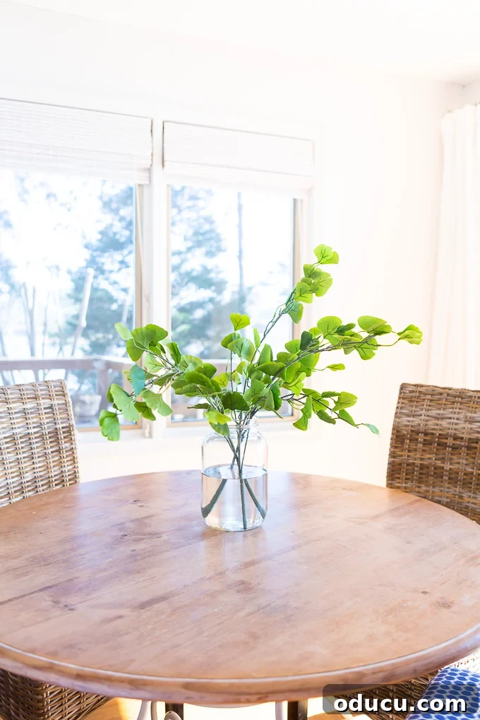 Large glass vase filled with water and fake house plant that is sitting on a round wood table.