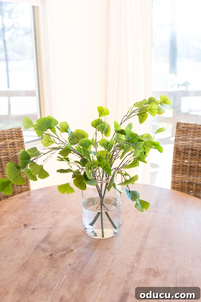 Fake Gingko house plant in large clear glass vase filled with water on top of a round wood table.