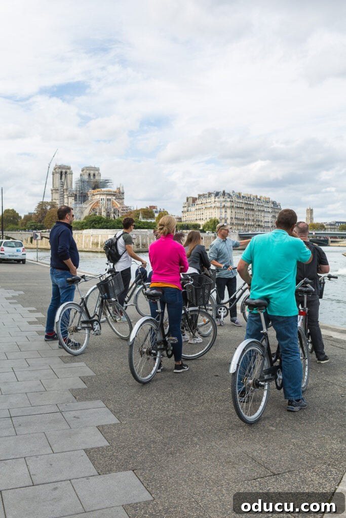 Bicycle riders on a tour through Paris pausing along the picturesque Seine River to listen attentively to their tour guide, with charming Parisian buildings in the background.