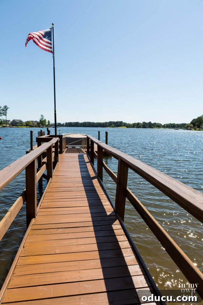 The Style Edit, Vol. 12 7 A long, beautifully stained lake dock extending into calm waters, with an American flag gently blowing in the breeze, evoking a peaceful scene of summer and reflection.