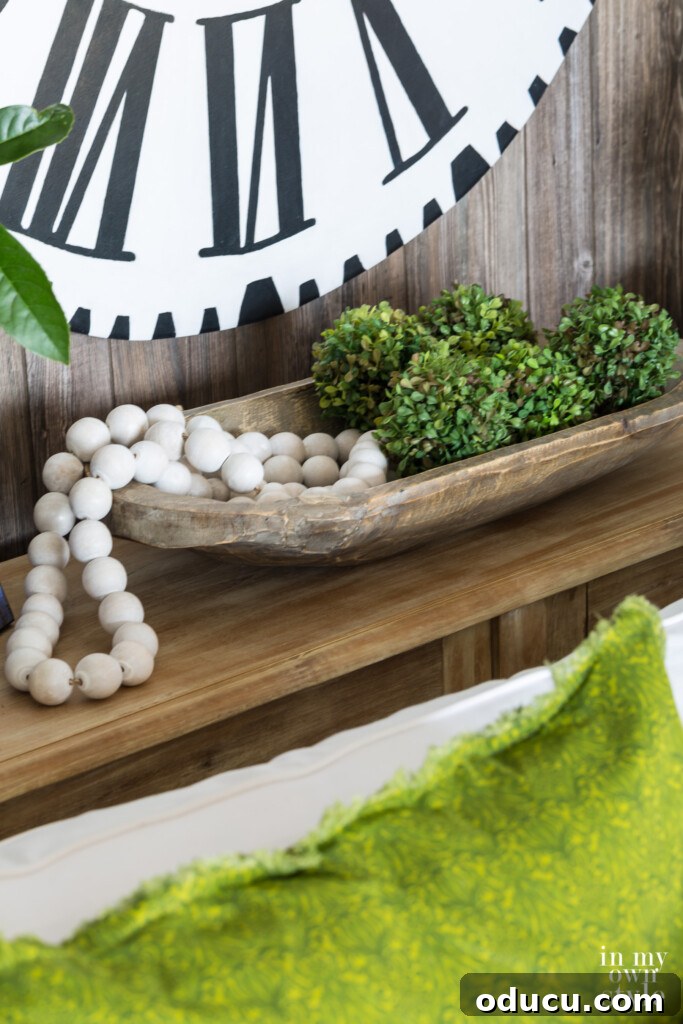 Effortless Summer Style 16 Dough bowl on sofa table filled with moss globes and wood beads. white clock with black Roman numerals on the wall.
