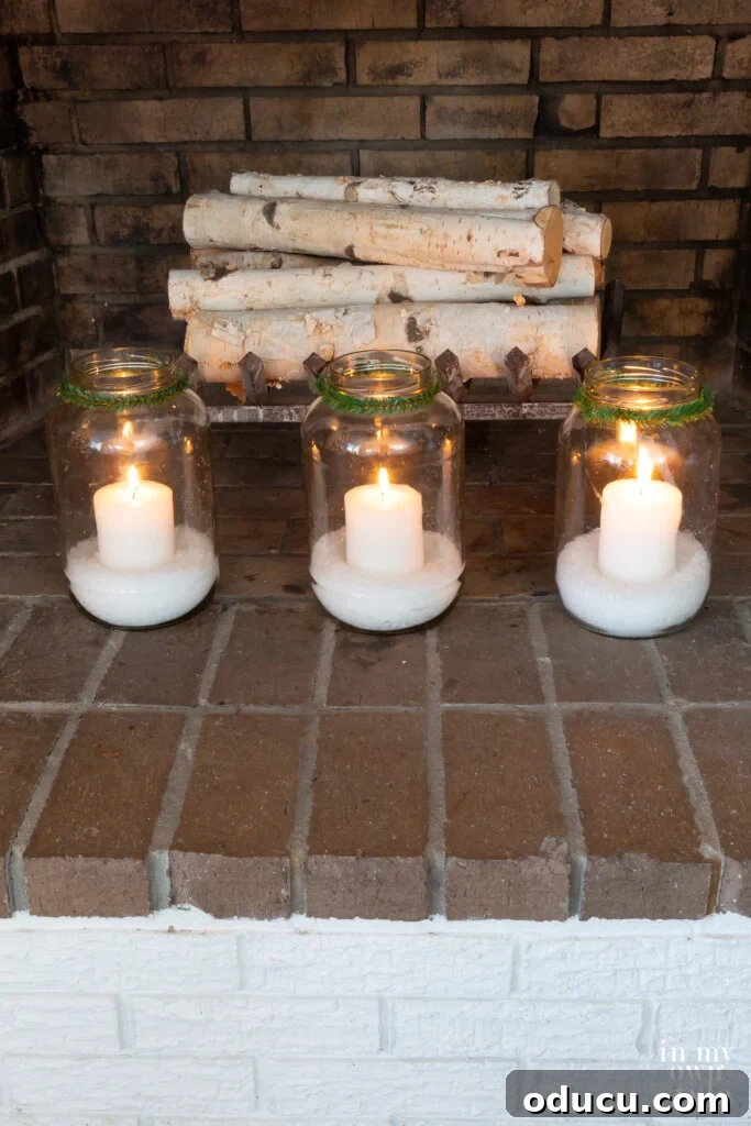 Three large glass jars filled with fake snow and white candles in a fireplace, creating a cozy winter scene.