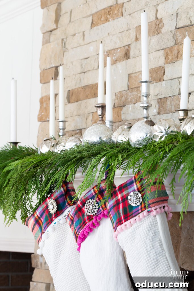 Three white Christmas stockings hanging on a cedar-draped fireplace mantel, illuminated by candles.