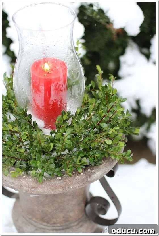 A red candle in a glass hurricane on an outdoor planter urn, adding a festive touch.