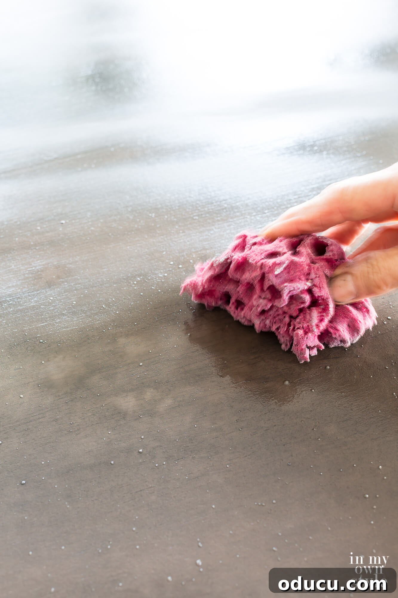 Zinc Effect Wooden Table Surfaces 13 A hand using a sea sponge to dab light grey, watered-down paint onto a surface, a key step in creating the mottled texture of a faux zinc finish. The technique demonstrates how layers are built for a realistic metal look.