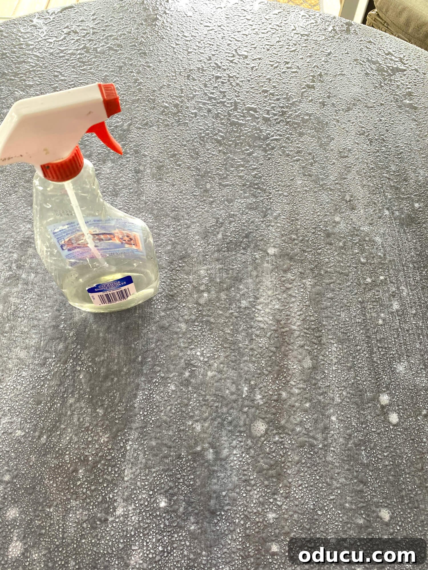 A hand spraying water over wet layers of paint on a tabletop, a technique used to soften and blend colors, creating a distressed and layered faux zinc finish. Mineral spirits might also be used in combination with water for specific effects.