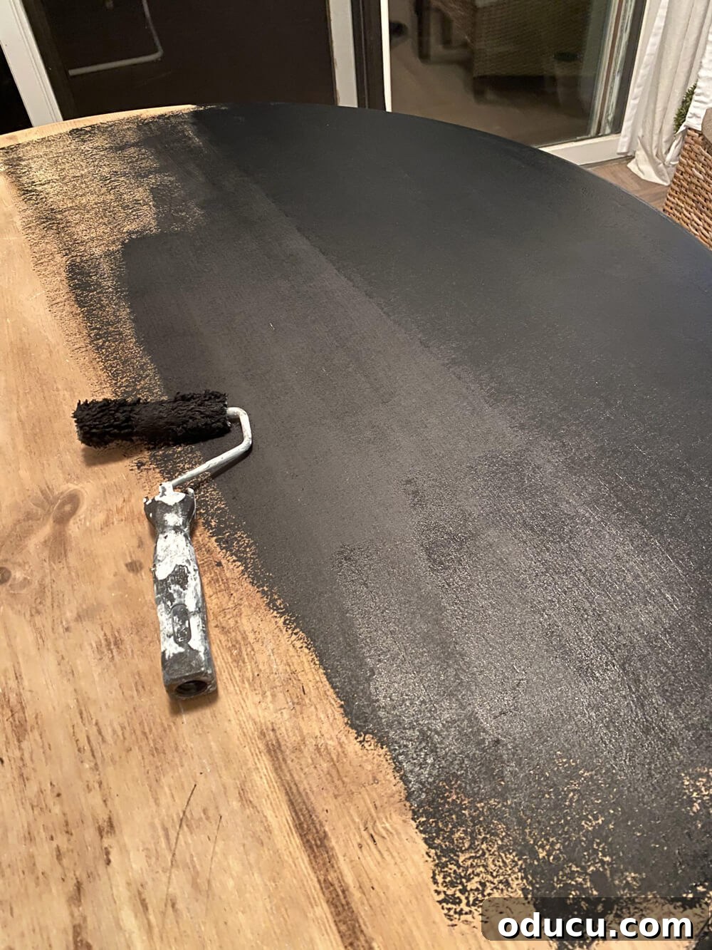 A round wood dining table in the process of being painted with a black base coat, demonstrating the initial layer essential for achieving a faux zinc or slate finish. The paint is applied evenly to cover the entire surface.