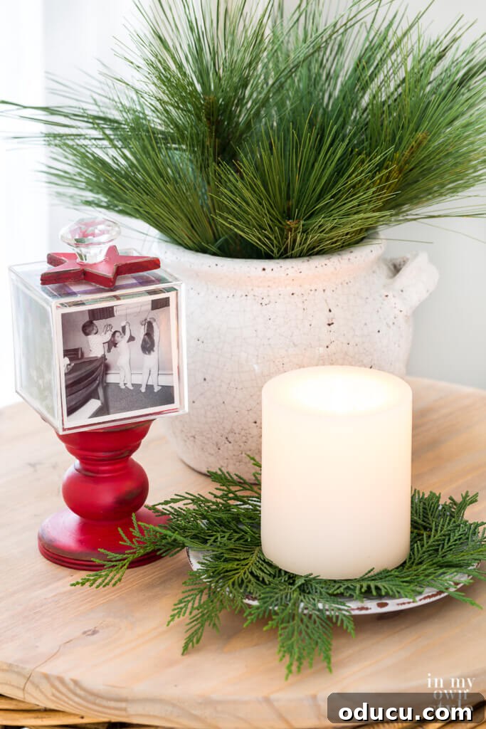 Effortless Festive Home Styling 15 A festive side table decorated for Christmas, featuring a red photo cube pedestal, a candle nestled on cedar sprigs, and a planter filled with pine, offering easy and affordable holiday styling ideas.