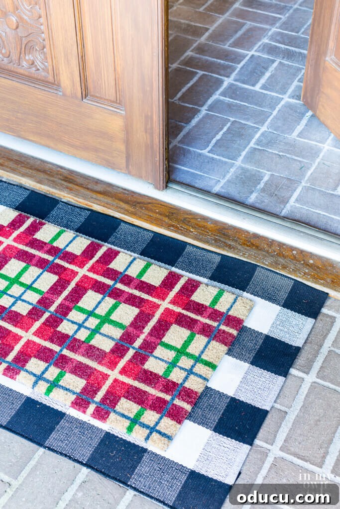 Effortless Festive Home Styling 3 A festive front porch entrance with a red and green plaid doormat layered over a black and white check mat, welcoming guests to a holiday-decorated home.