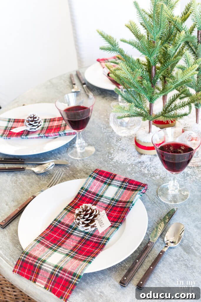 Effortless Festive Home Styling 26 A close-up of a charming Christmas table setting, featuring a rustic red plaid theme, artificial snow, and elegant pinecones with painted white tips serving as unique place card holders.
